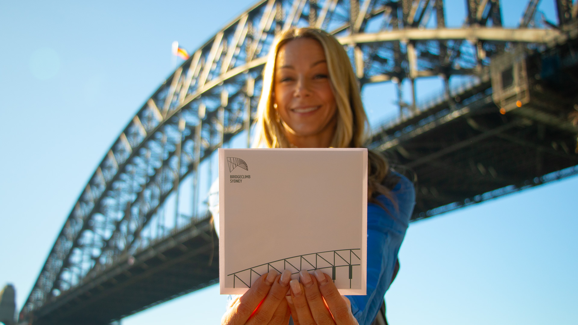 blonde haired woman in a bridgeclimb suit stands holding a white envelope with the bridgeclimb logo with the sydney harbour bridge in the background