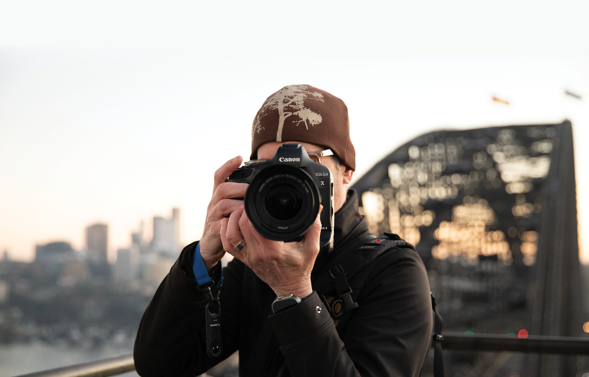 man holding a camera in front of his face taking a photo with the sydney harbour bridge in the background