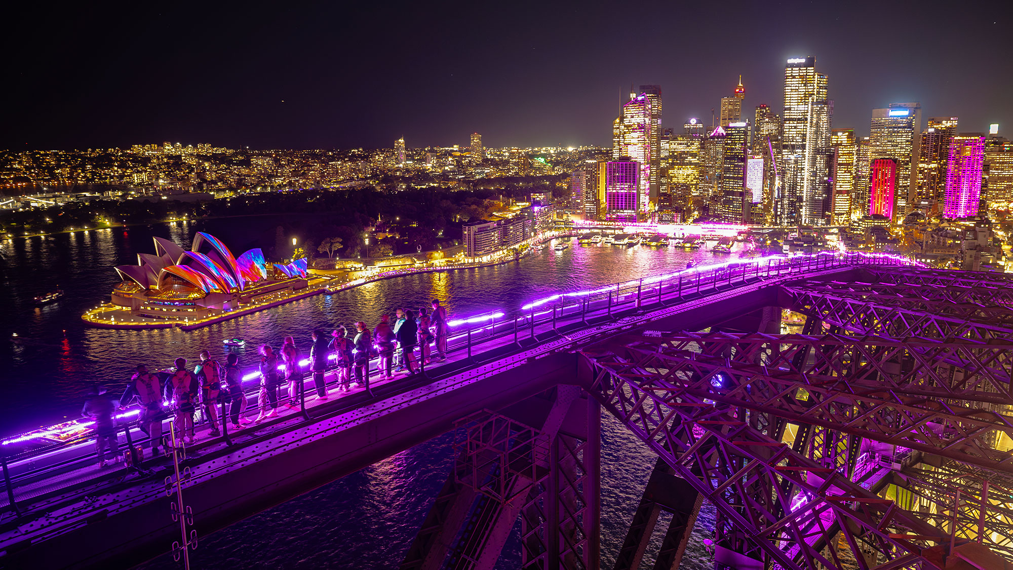 climbers walking along the iconic arches of the sydney harbour bridge with a view of the harbour and the sydney opera house during the vivid sydney festival