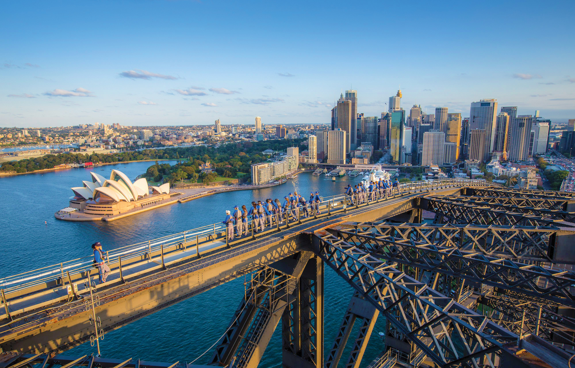 climbers walking along the iconic arches of the sydney harbour bridge with a view of the harbour, the sydney opera house and city skyline in the background