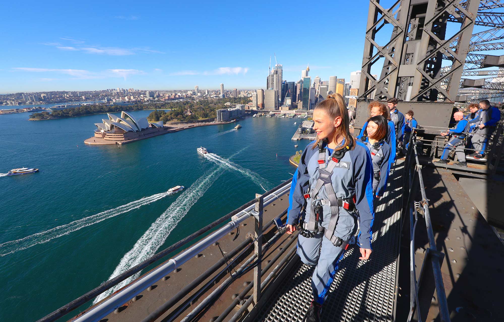 Blonde haired woman climbs the lower arch of the Sydney Harbour Bridge and smiling while enjoying the view of the harbour below including the Sydney Opera House